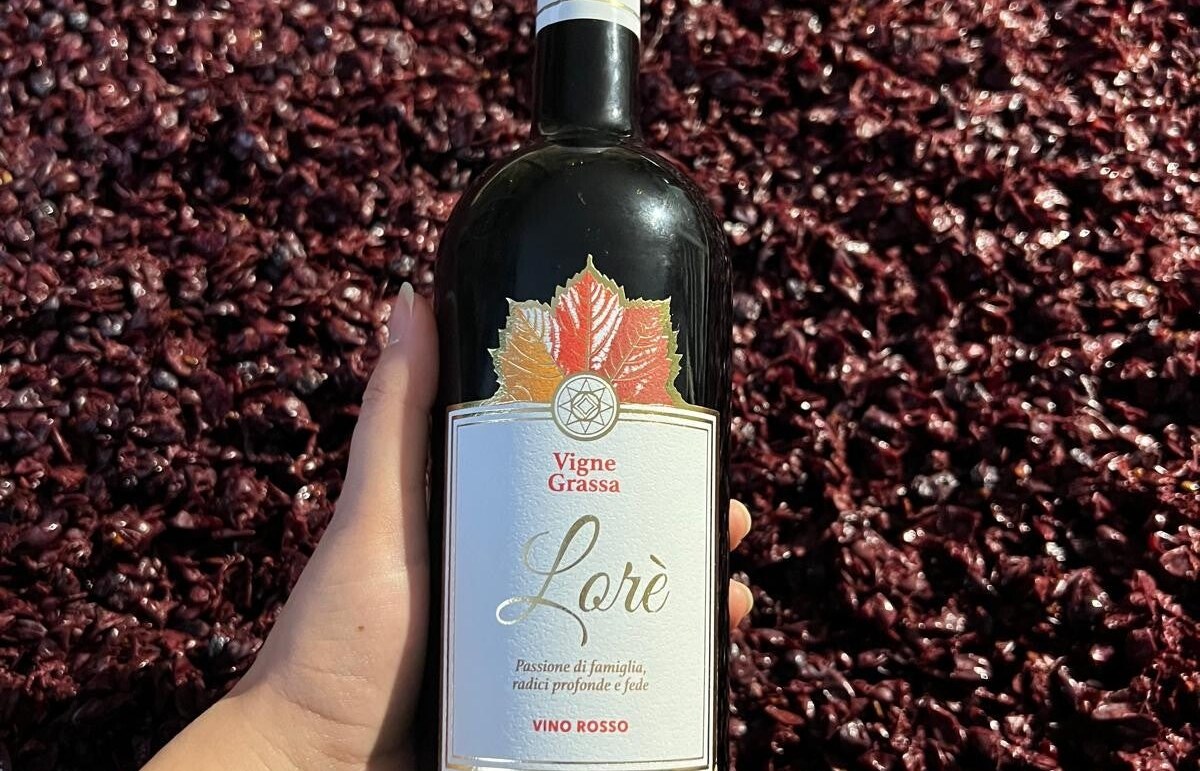 Mano che regge una bottiglia di Lorè Vino Rosso Vigne Grassa con foglia di vite sull'etichetta, sopra vinacce rosso scuro